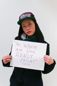 Asian woman rolling her eyes as she holds a sign saying, "No, where are you really from?"