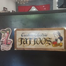 1950s tattoo artwork displayed at a black counter. Shows a cobra saying 'Cash only.' The larger sign shows a cartoon woman with a banner that reads, "Trixie," and the larger lettering: Custom Electric Tattoos. AtBroadway Tattoo, Sydney
