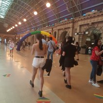 People walk and talk at the grand Central Station, Sydney. There is an arch of balloons made in the pride flag colours