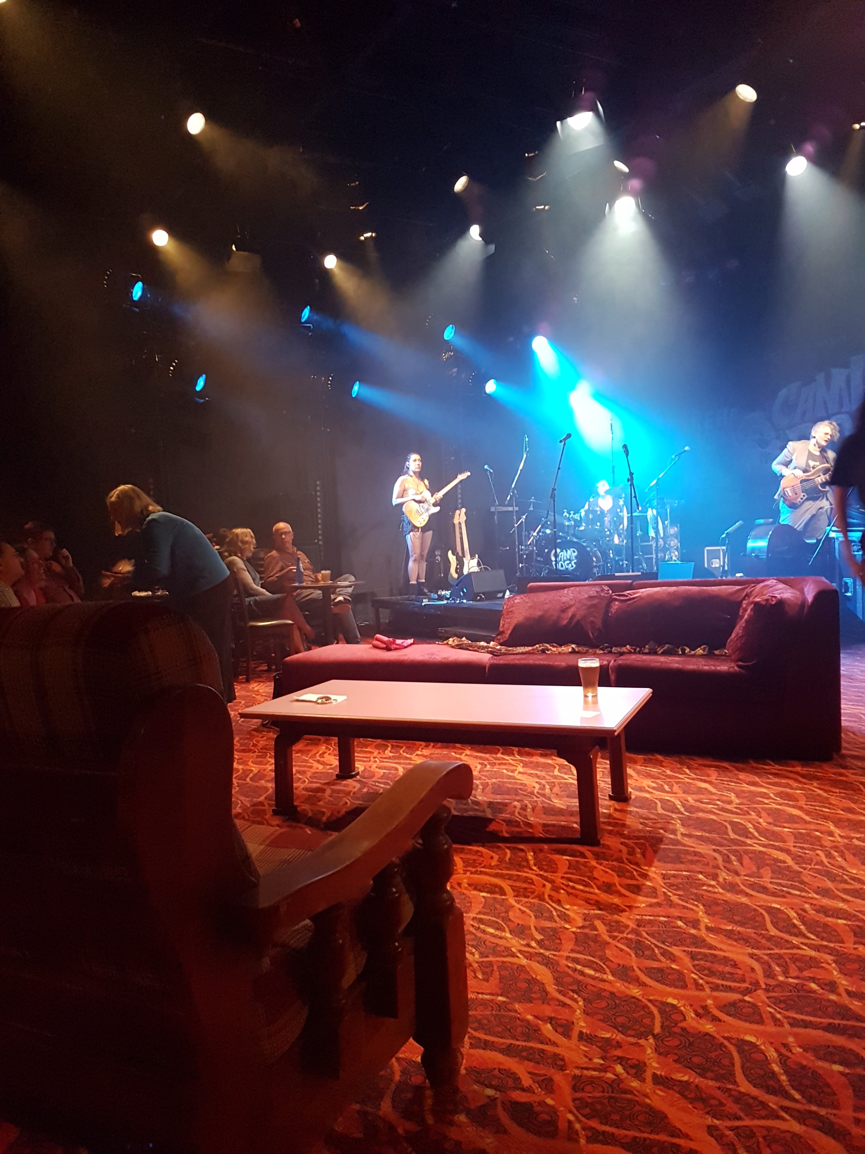 An open stage is set up to look like a lounge room, with velvet couches and large chairs. A beer sits on top of a coffee table. On the side background, the audience can be seen looking to the stage. In the far background, a woman is bent over talking to the audience, while band members are practising on their guitars onstage