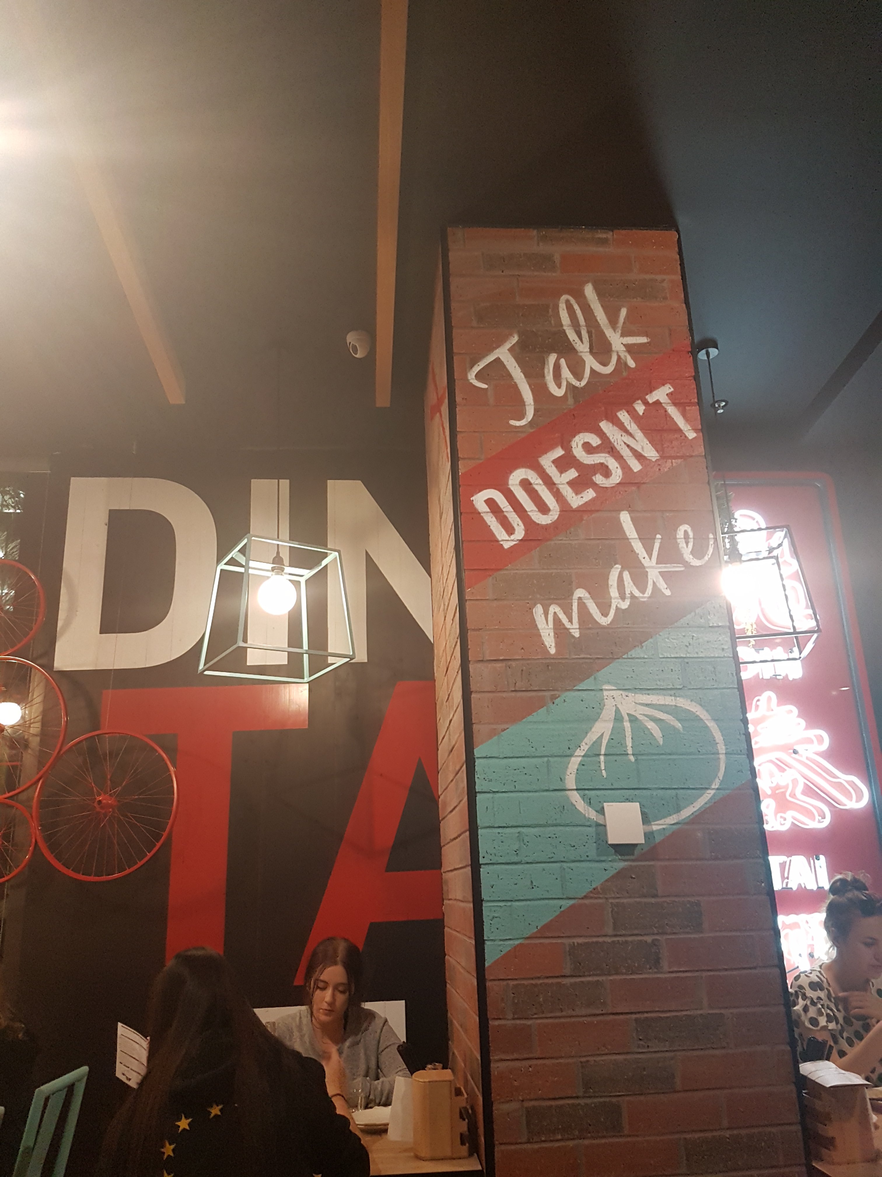 Indoor column of Din Tai Fung restaurant with the words 'Talk doesn't make' and a drawing of a large dumpling. White women are sitting on either side of the sign, looking down at their tables