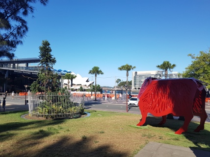 A red metal merino sheep stands at 2.5 meters, in the mid-ground, surrounded by trees, near the Oversas Passanger Terminar, and the Sydney Opera House in the far background