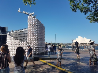 People in the foreground take photos of a large white sculpture, which on closer inspection is made of hundreds of white pig faces with pink noses and ears, and in other places, pig bums with pink curly tails. The sculpture is immersive, and you can walk around the inside in a circle. The Sydney Opera House can be seen in the background on a sunny day